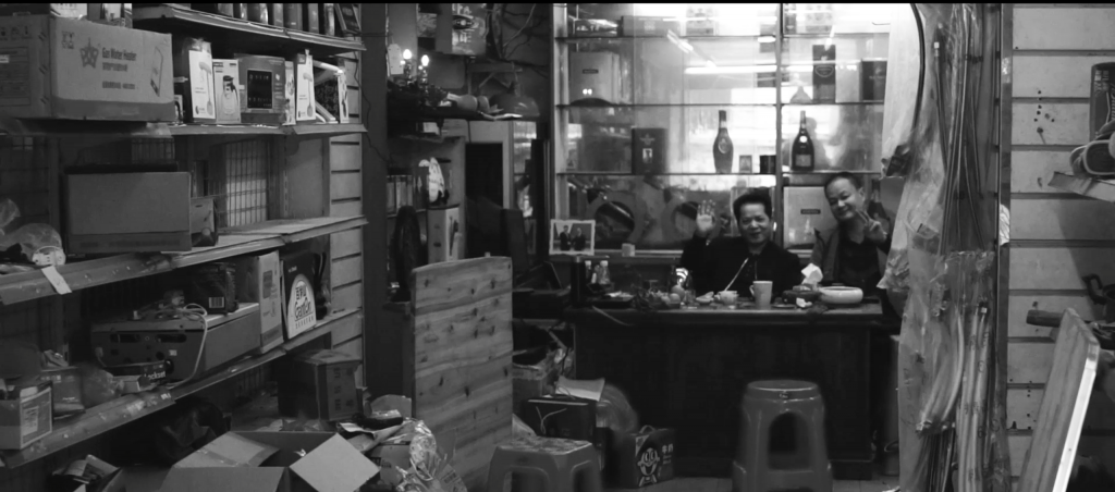 Black and white image of two men sitting inside a shop cluttered with objects, both are smiling and waving to camera.