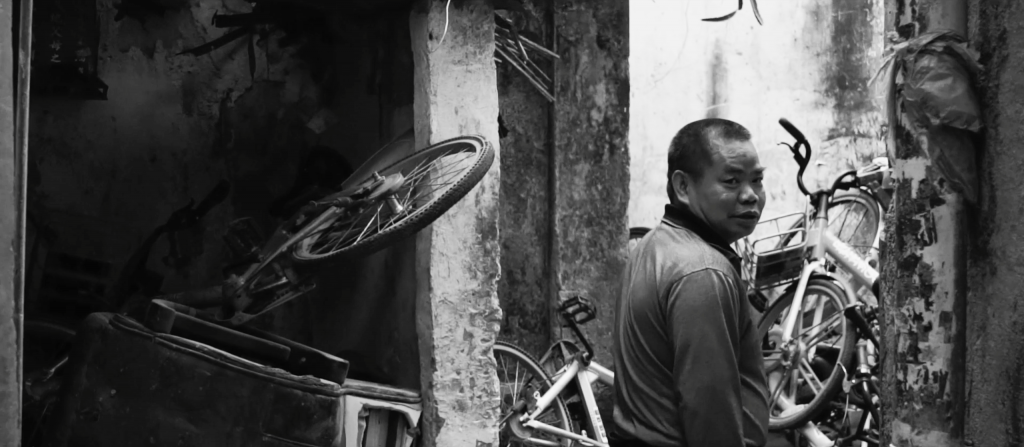 black and white image of a man in an alleyway, turning to look over his shoulder towards the camera. In front of him are many 'pay to use' bikes piled up high.