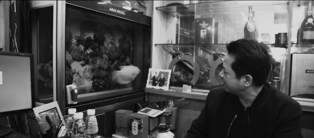 Black and white image, man looks to his left to face his aquarium which has a fish inside, floating next to a photograph of the Chinese leader Xi Jinping