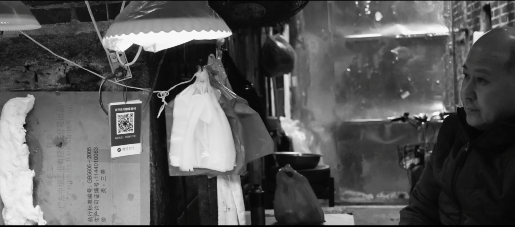 Black and white image of man looks to the right, at his outdoor butchers stall. Low level hanging lights and plastic bags are in the frame. He is wearing a warm coat suggesting that it is a cold day.