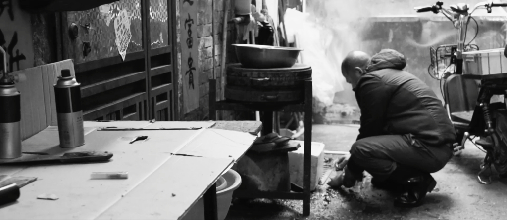 Black and white image of a man crouching down to cut up a piece of pork. There is a table with cardboard on the top which serves as the butchers main selling platform.