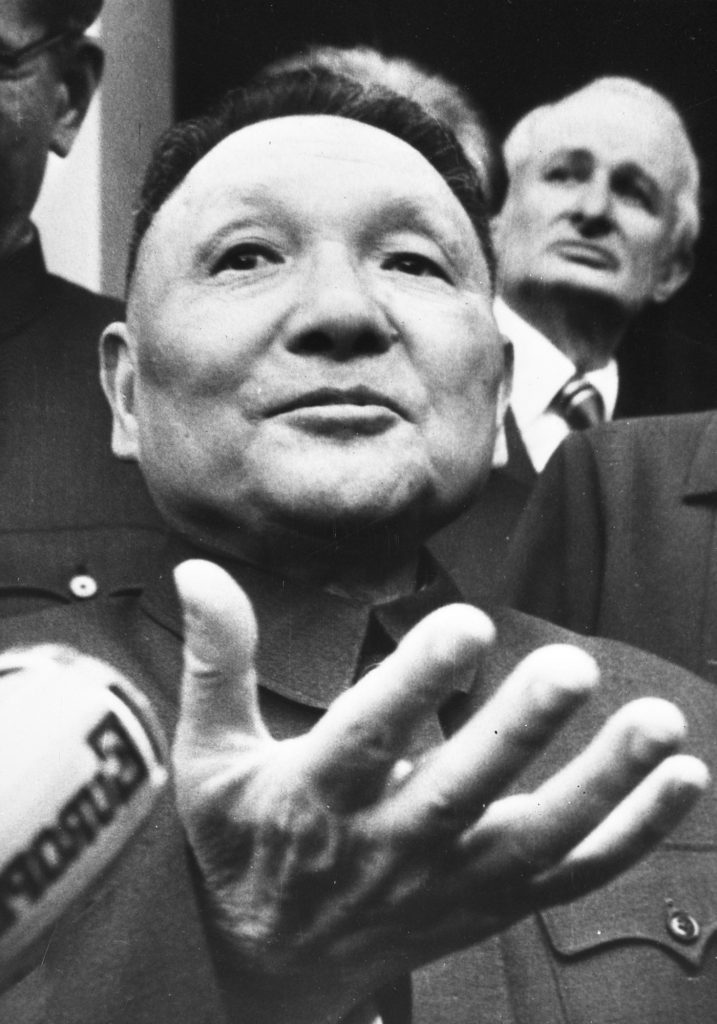 Black and white image of Deng Xiaoping in mid-speech, raising his right hand as he addresses and audience in Paris (1976)