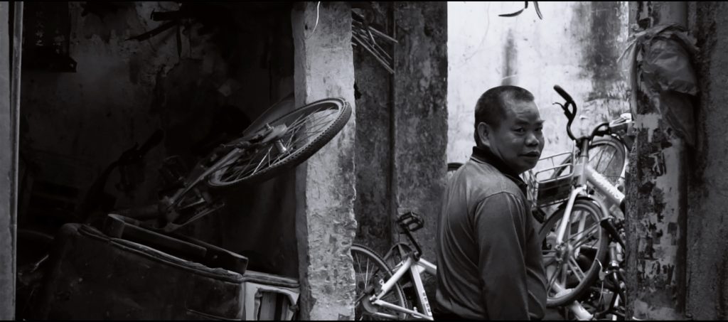 Black and white photograph, with a man to the right of the image, looking out at the viewer from the photo. Several (what appear to be) abandoned bicycles are piled on the corners to the left and right of the image.