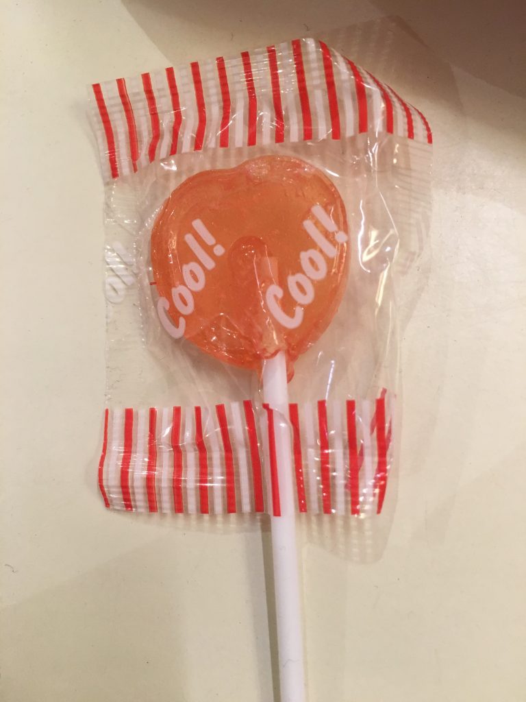 An orange heart-shaped lollipop wit the words 'cool!, cool!' written on the wrapper.