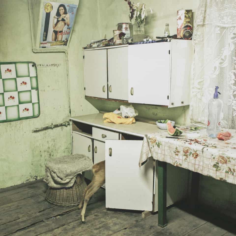 A photo of a kitchen with white cabinets. A pin-up girl is on the wall and a table nearby has a tablecloth. The butt of a little fawn is sticking out of one of the open cabinet doors.