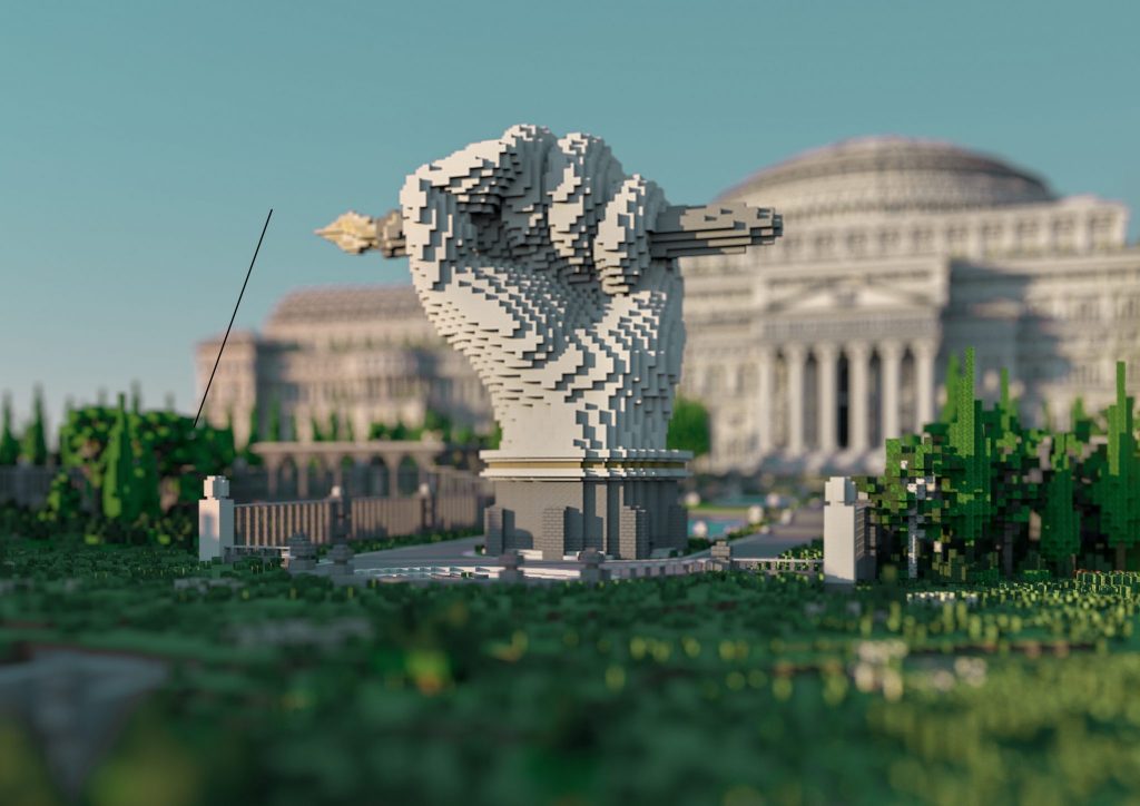 An image from The Uncensored Library project. This is a view of the front entrance of the virtual library in Minecraft. A virtual sculpture of a raised fist holding a dip pen in a garden with trees and grass is in the foreground. The library itself is slightly of focus in the background in order to emphasize the sculpture. The library is built using a neoclassical architectural style with big columns and a domed rooftop. It is intended to resemble well-established institutions such as the New York Public Library, as well as stylistically allude to the authoritarian structures the project aims to subvert. 