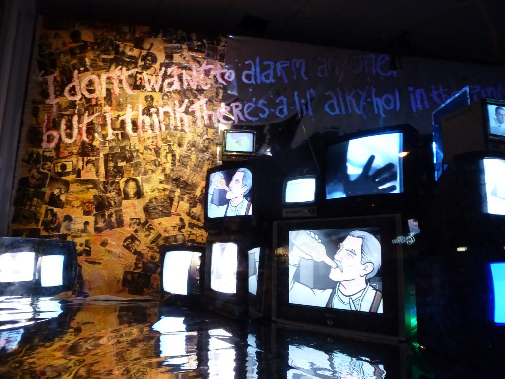 Installation view of an exhibition. On the left of the image is a tree-shaped collage of many, many black and white images (it's not very clear as the images are small, but some are portraits of people) - they are lit in a sunshine yellow light. The floor of what is assumed to be a gallery is chrome, and reflects the rest of the room. 12 TV monitors make a wall on the right of the image, on 2 screens are an animation of a man drinking from a bottle, on another is a hand touching the screen, other screens are whited out or unclear. On a On the wall and written over the photos in large handwriting is written: I don't want to alarm anyone but I think there's a lil alkyhol in the..." unreadable from there.