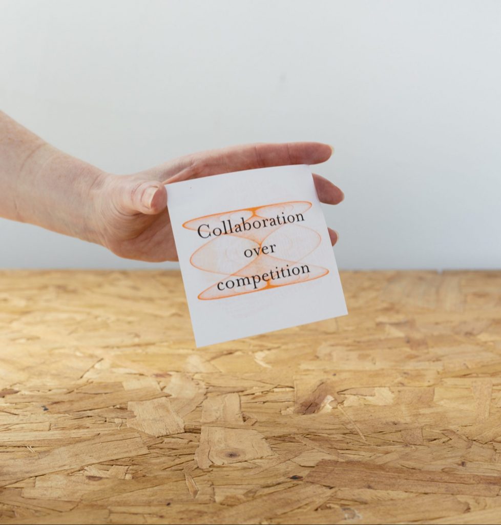 Hand holding a white card up to the camera. Text on the card is superimposed over an orange helix. It reads "Collaboration over competition".