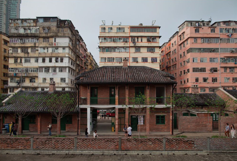 Photograph showing the entrance to Cattle Depot in Hong Kong. The entrance is an archway inside a red brick building on two storeys, with two smaller outbuildings either side. The roofs of the buildings are covered in a dark rough tile, which is in parts covered in a red moss. Behind the buildings are tenement buildings, each approximately 10 storeys high; the one on the left is pale blue and grey, the central one is cream with orange stripes running beneath the windows, and the one on the right is pale salmon-orange/pink. The sky is overcast, pale grey-blue.