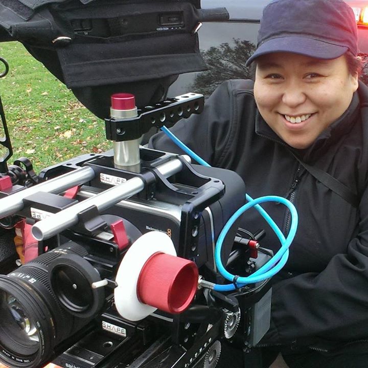 Photo of the artist holding smiling behind an industry film camera. 