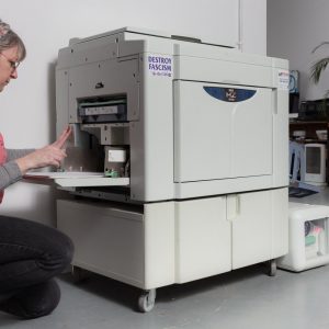 Damien Robinson, a white woman in a pink t-shirt, adjusting the paper feed in a risograph machine.