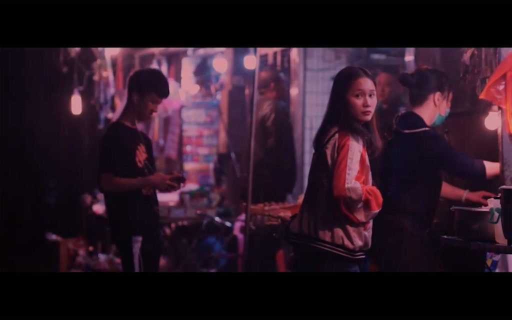 Colour photo, with red, blue, purple hues. A young woman looks to camera, whilst a street vendor prepares a meal for her.