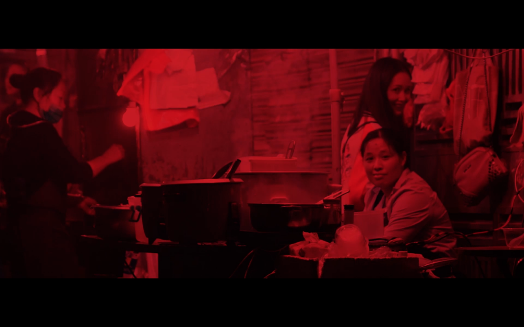 Colour photo in red and black hues. A young woman and street vendor looks to camera smiling, another street vendor prepares the young woman's food. Plastic bags are strung up, pos and pans are steaming with food.