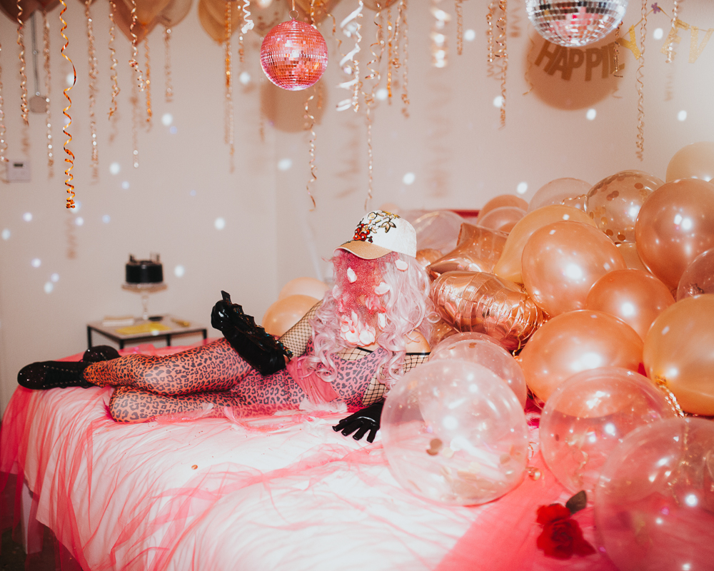 A person lies on a bed wearing pink and black leopard skin leotard and leggings, and black fishnet top beneath, also wears black gloves and boots. They have a pink wig and cap on, their face is covered by tight dark pink curls falling from beneath the visor of the cap. 
Metallic bronze and clear balloons are piled on the right side of the bed and hang in the air. Golden twisted ribbons hang from the balloons. 2 disco balls also hang from the ceiling. A red chiffon sheet covers a white bedsheet, which the person lies upon. 