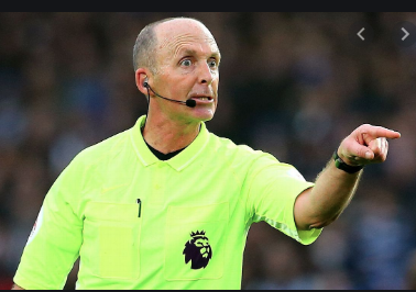 A front facing photo of a football from the waist up, wearing a neon tshirt with a small black lion logo, he has a Britney or call centre style microphone attached to his face, he is balding with some grey short hair around the edge. His face is tensed and wide eyed and his left arm is outstretched as he angrily points at something or someone not visible in the picture. In the background is a very soft focus football crowd.