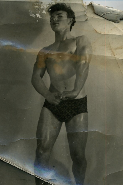 A photo of an old printed photo which is very crumpled and worn, with fold marks and a dogeared corner. The printed photo is from a front perspective and in black and white. Centre is a teenage man standing, visible from the ankles upwards. He is wearing only a pair of tight black pants, he is posing in a bodybuilder stance, with his fingers hooked together and his arms turned outwards, to show the extent of his arm muscles, his skin is slightly shiny as though it has been oiled. His head is tipped upwards and he looks off into the distance. He has a fluffy dark head of hair in a messy quiff style. He looks calm and confident. 