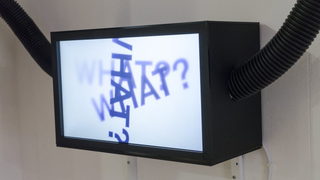 A black rectangular lightbox with a white screen has the word what with a question mark written 3 times on it, black pipes leave the box on the left and right sides.