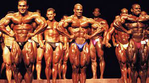 Several rows of men on a stage at a bodybuilding competition. They are all in tiny tight pants with bulging muscles all over the bodies. They are very oily and shiny. The front row of three men have their hands on the hips in fists to accentuate the muscles in their torso and arms. The two on the left and middle are smiling, the one on the right has his cheeks puffed out as if he is straining. The rows behind are stood more casually waiting their turn. The layers of bodies and muscles in the picture blend together making it difficult to differentiate between each person. Their backdrop is plain black. 