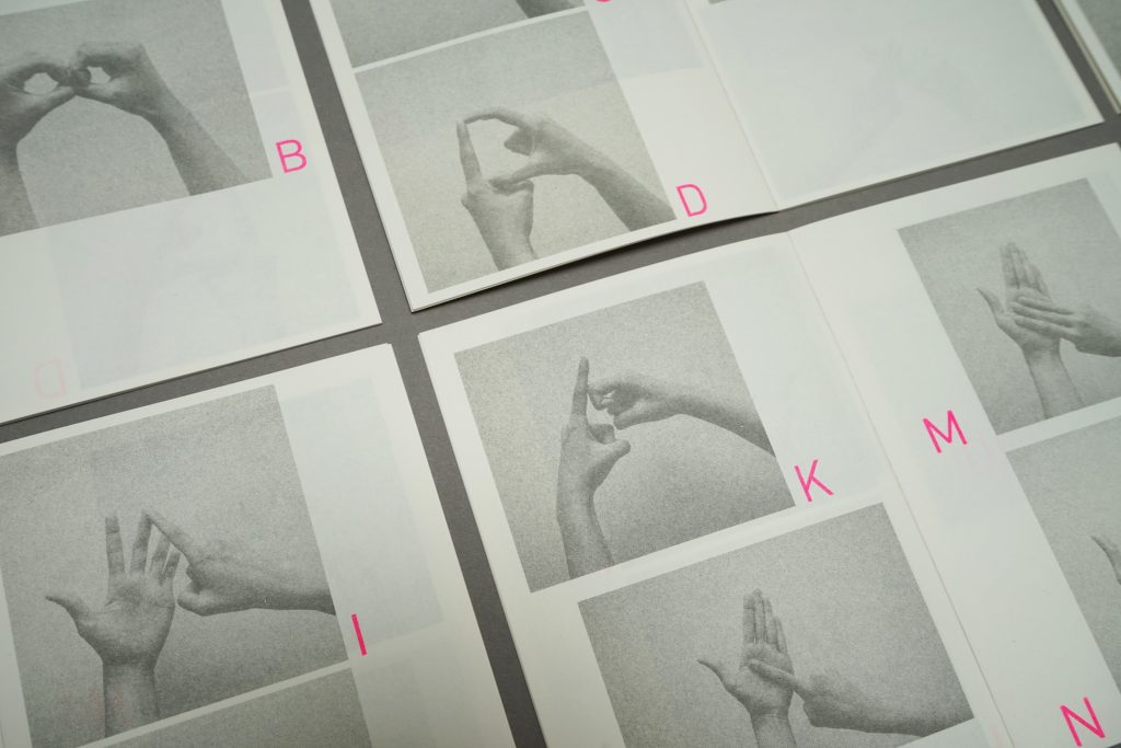 Documentation image of four BSL booklets that is shown with its pages open laying each other- all images of hands are grey-scaled, showing bright pink alphabets of D, K, M, N, I, B in clockwise direction.