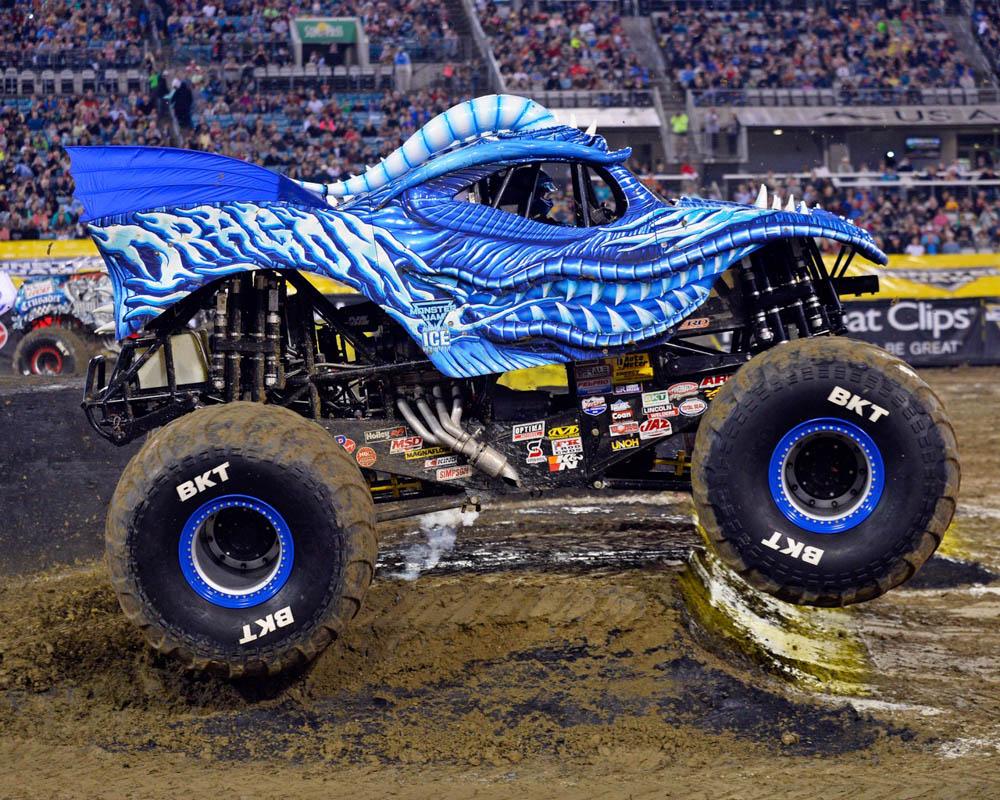 Dragon Ice monster truck, a compact monster truck with a blue dragon body design with scales and teeth and spikes and big wheels. There are colourful logo stickers on the black undercarriage. It is doing a small jump over a very muddy ramp in a stadium filled with people in the background. 