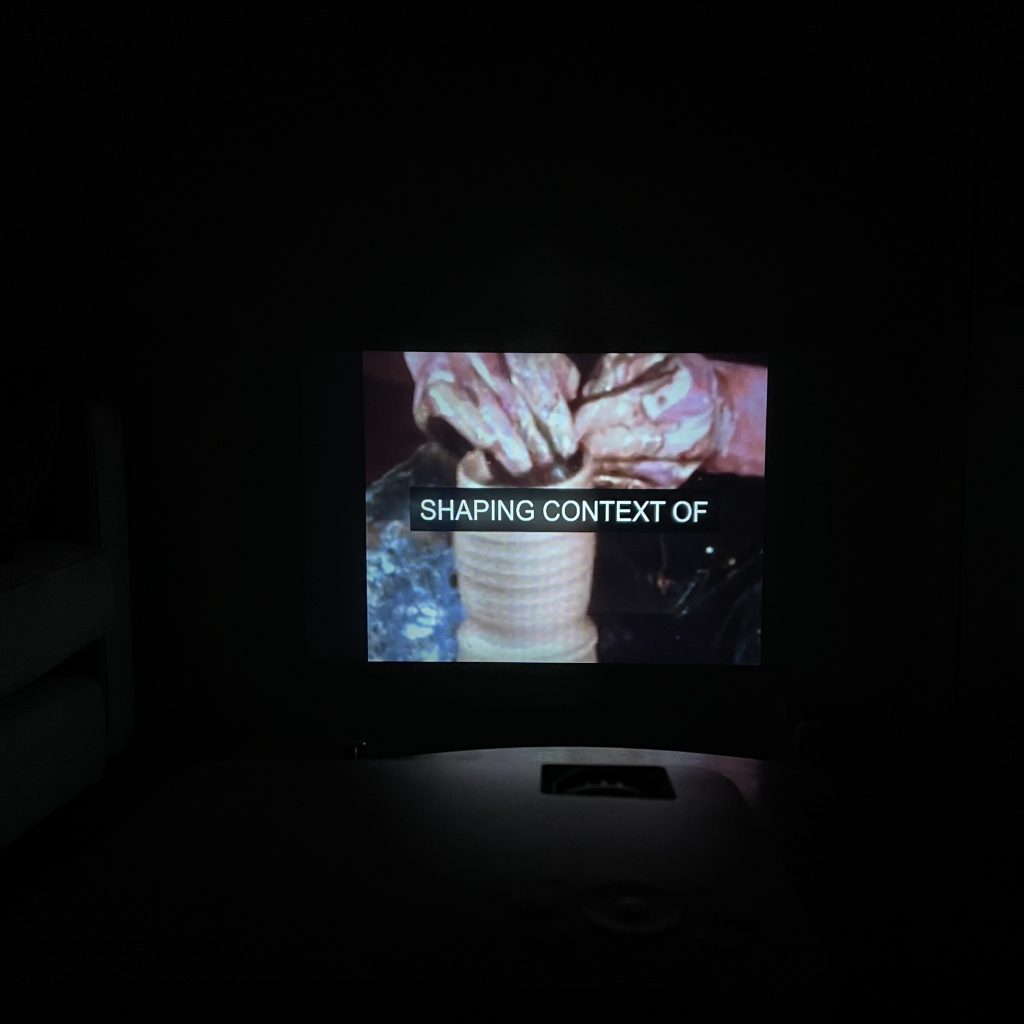 A picture of a projection on a wall. The picture is very dark as it was taken in a room without lights on. A pair of hands are shaping clay into round cylinder shape. There is a white subtitle against black background in the centre of the image that reads "SHAPING CONTEXT OF" Just below the projected image, there is a hint of the shape of the projector itself.