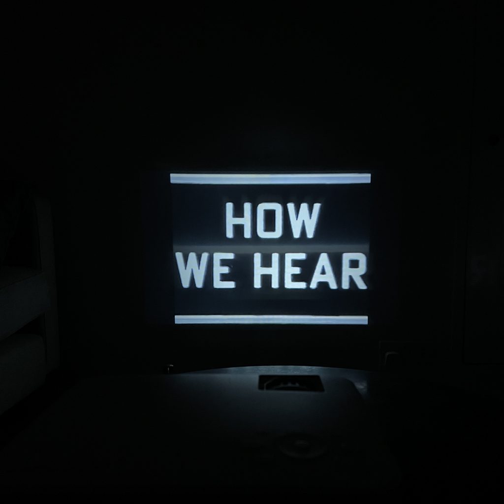 A picture of a projection on a wall. The picture is very dark as it was taken in a room without lights on. The words shown in the projected image  are displayed in two lines of HOW and WE HEAR. The text has a thick white horizontal line above and below it. Just below the projected image, there is a hint of the shape of the projector itself.