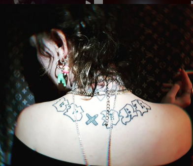 An image of the back of my head and the top of my back, a tattoo which says 'BB X PNK' which is short for baby punk, a performance persona of mine. It is in black old fashioned font and the 'X' is made of bullets. 