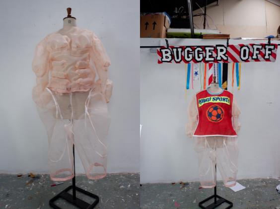 A split-screen image of two headless torso mannequins adorned with fleshy mesh-like skin. The left mannequin has the same perspective as a changing room mirror and bulging muscles made of fabric. The right mannequin is posing for its birthday snap beneath a striped red and white banner that says "bugger off" in all caps. Multicoloured tassels hang from the centre of the banner and the mannequin is wearing a red bib that has a neon orange football with hard sports written above.