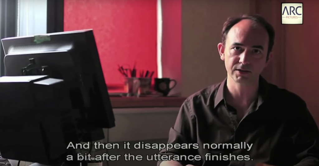 Screen capture from Youtube The Invisible Subtitler- a person is sitting on right, wearing a dark coloured shirt. There is white subtitle at bottom saying "And then it disappears normally a bit after the utterance finishes". On left there's a black computer screen monitor facing the person, showing only backside of it. The background has red tone, coming from red coloured blind in the window. At the bottom of window, there is 2 cups of utensils and a empty mug cup. 