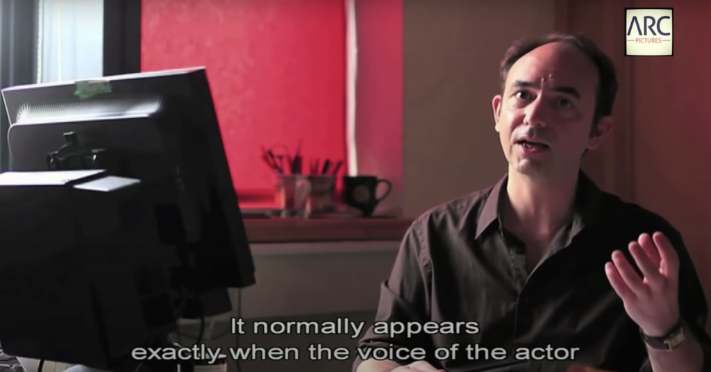 Screen capture from Youtube The Invisible Subtitler - a person is sitting in the right, wearing a dark coloured shirt. The white subtitle at the bottom says "It normally appears exactly when the voice of the actor". On left there's a black computer screen monitor facing the person, showing only backside of it. The background has red tone, coming from red coloured blind in the window. At the bottom of window, there is 2 cups of utensils and a empty mug cup. 