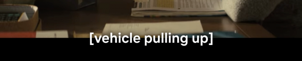 Cropped image of film only showing the subtitle that says [Vehicle pulling up]. Behind the subtitle you can see cropped image of wooden table with paper stacks on it.