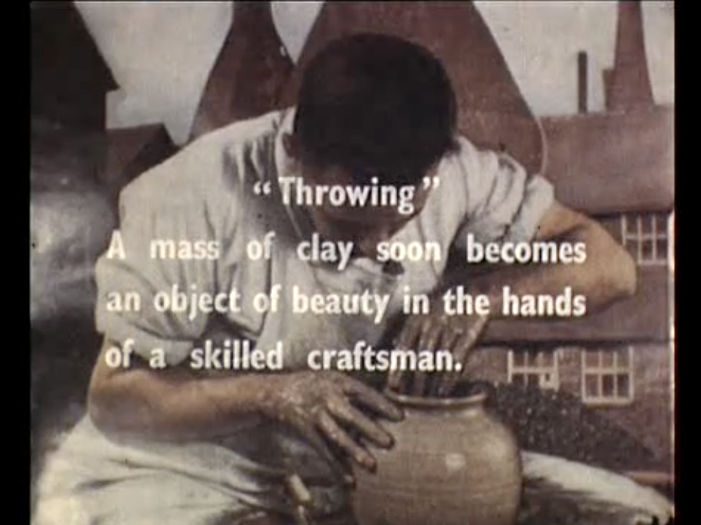 Scene from Sam Hanna's film "Village Potter"- the person who is wearing white shirt and white apron is throwing a round shape of pottery. There is description of what "Throwing" means - "A mass of clay soon becomes an object of beauty in the hands of a skilled craftsman" in simple white font located in centre of image. Behind the person, there is an image of a brown house with small white framed windows and pointed roof.