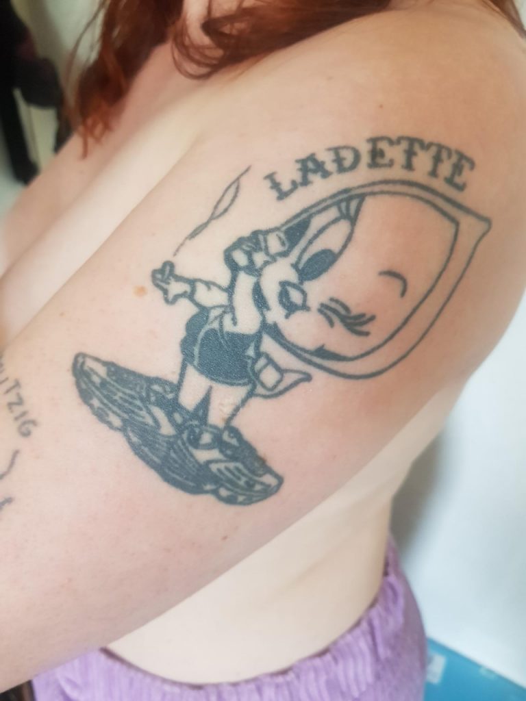 A close up image of the side of my arm. On it is a medium sized tattoo of Tweety Bird, a cartoon bird from Looney Tunes. It is a black outline tattoo. Tweety is wearing a hoody and large trainers, in their left hand they are holding a tinny and in their right hand a smoking cigarette. They are winking their left eye. Over the top of their hooded head it says 'LADETTE' in old school tattoo style writing. 