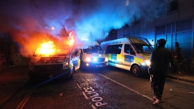 three British police vans parked in a narrow street at night. They are parked at angles and not in a straight line. The left van is on fire which is bursting out of the front window and sides. The bonnet is covered in graffiti which says 'kill' and 'ACAB'. Smoke is billowing and filling the top part of the image, it is glowing orange and red from the fire and blue from the van's lights. The vans in the middle and on the right are not on fire. Their headlights and blue emergency lights are on. A silhouette figure of a person is visible in the foreground on the right from behind. 