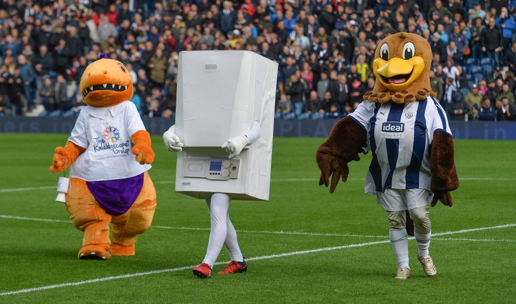 A green football field with busy stalls filled with fans in the background. In the forefront is three mascots walking together, on the left is an orange dinosoar with a white shirt on holding a charity collection tin, in the middle is a white boiler with white arms and legs and red football trainers, on the right is an eagle with a white and navy blue striped football kit on. 