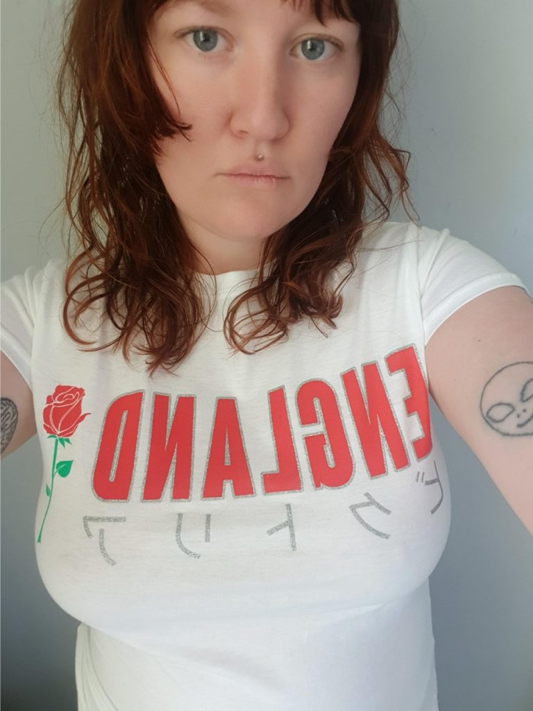 A selfie I took this morning wearing my England  tshirt. The perspective is  from eyebrows to stomach. My face is tired and expressionless and my eyes a bit watery because my baby has been waking me every hour during the night for a week. My hair is unsurprisingly messy and unwashed, hanging wavy at shoulder length. I have blue eyes and a small piercing above my top lip. The shirt is white and a cheap almost see through material, the sleeves are capped and my arms are showing slightly with some messy black outline tattoos, a smiling alien on the right and a brain on the left. On the chest of the tshirt it says 'ENGLAND' in red capitalised slim bold letters with silver glitter outline. The lettering is backwards because the phone has mirrored the image. On the left of the lettering is a red rose with a green stalk and leaves. Underneath is some silver glitter lettering in what looks like Japanese letters. I don't think it's a translation of ENgland because I looked it up and it looked different so I have no idea what it says.  