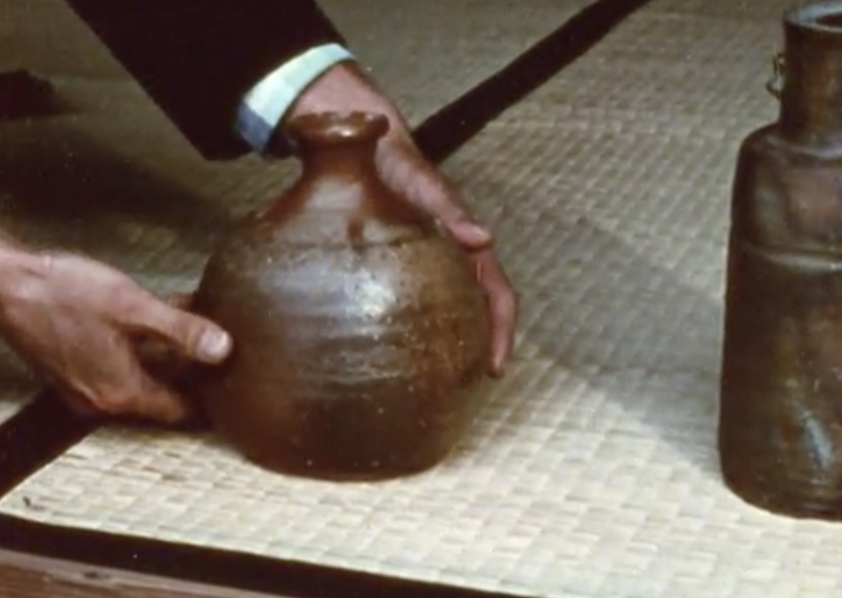 The hands of a Japanese Potter holding a brown glazed round ceramic piece. At the bottom of ceramic, there lays a yellowish white coloured bamboo mat that are lined with black lines. On the right there's another brown glazed ceramic that is long cylinder shaped with long neck. 