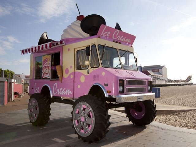 A pink and cream ice cream monster truck with large oversized wheels and a big soft white whip on top with a cherry. 