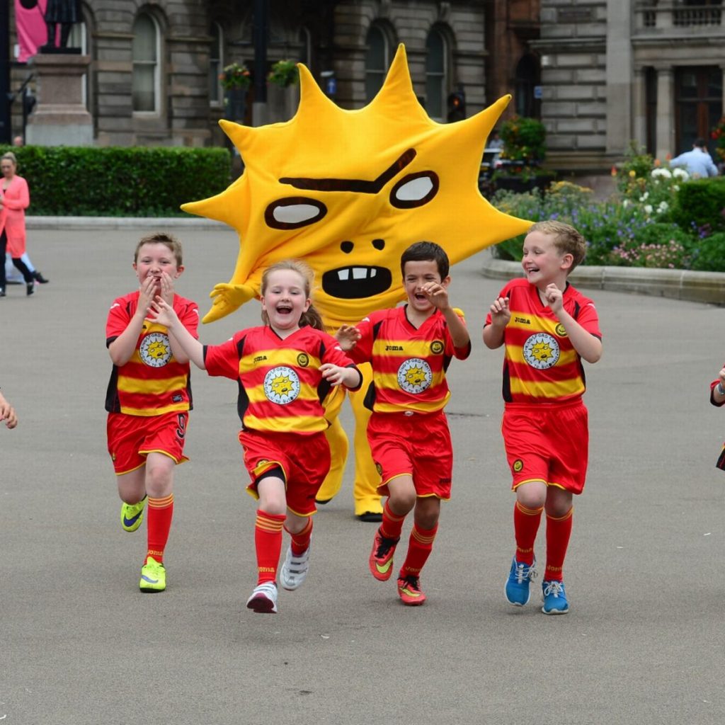 A town square is visible in the background with some planting beds and hedges. In the forefront is a person dressed in yellow trousers and a large mascot top which looks like a giant sunburst with a slightly angry cartoon like facial expression. They are chasing four children who look like they are around eight or nine years old, they look excited and slightly scared to be chased at the same time. They are all wearing red and yellow Partick Thistle football kits. Their arms are flailing up in excitement as they run. 