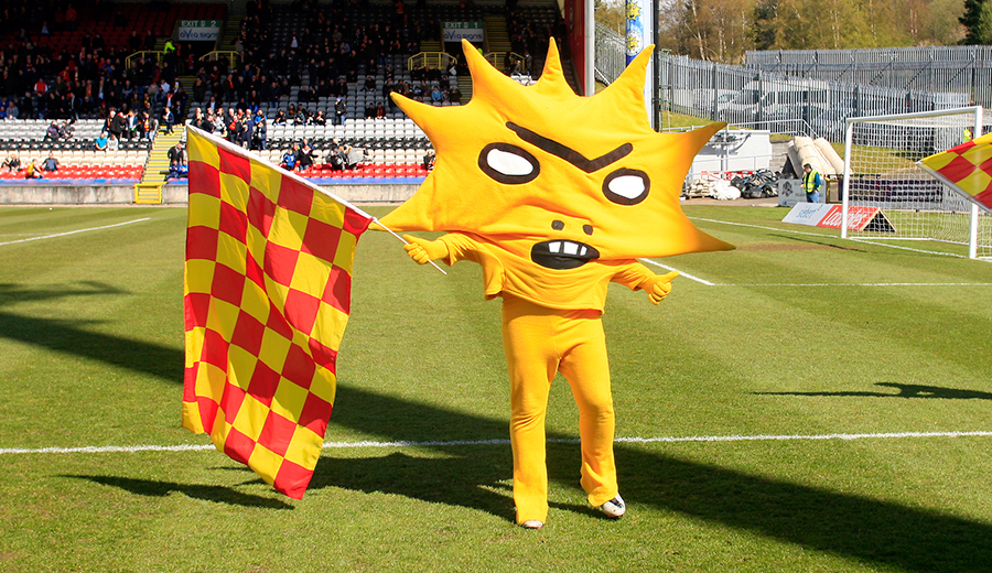 A green football field with a goal net and some fans in the stalls visible in the background. In the forefront is a person dressed in yellow trousers and a large mascot top which looks like a giant sunburst with a slightly angry cartoon like facial expression. It is holding a large red and yellow chequered flag and is giving the thumbs up with it's other hand. 