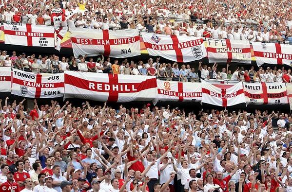 A sea of footy fans, almost all of them have their arms raised up and almost all of them are wearing red and white footy outfits. Many have large England flags which are a white rectangle with a red cross which they have added their hometown names, their own names or their own slogans to such as 'Tommo', 'Bollocks' and 'Redhill', which they have hung off the edge of the stands. 