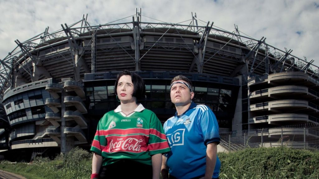 Two people stood outside of a large footy stadium shot slightly from below so that the stadium looms over them like an ominous space ship. It's shadowy in front of a cloudy sky. The two people are brightly lit and gaze upwards, both visible from the waist up. The person on the left has a black bob haircut and they are wearing a green and red footy shirt. The person on the right has short brown hair and head band across their forehead and a blue footy shirt with white details. They both have their hands in their pockets, they look slightly pensive like they have seen something in the sky that is about fall down in front of them. 