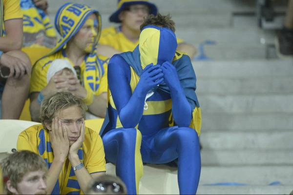 Football fans in blue and yellow sat in the stands of the stadium. Central is someone in a full body blue and yellow morph suit with a flag wrapped around their neck with their head in the hands, all the fans around them look sad and fed up too/ 