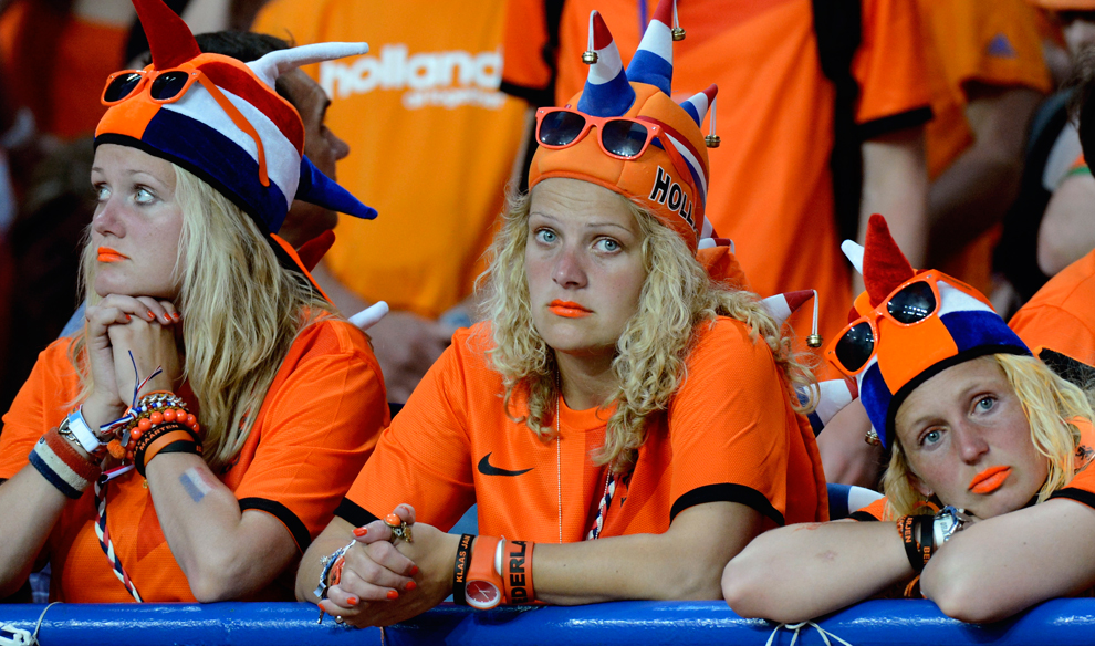 Three football fans all dressed in orange with spiky orange, blue and white hats on with 'Holland' written on them, orange sunglasses on their heads and orange bracelets and lipsticks, leaning on a blue pole at the edge of the stand in the stadium looking sad and fed up. 