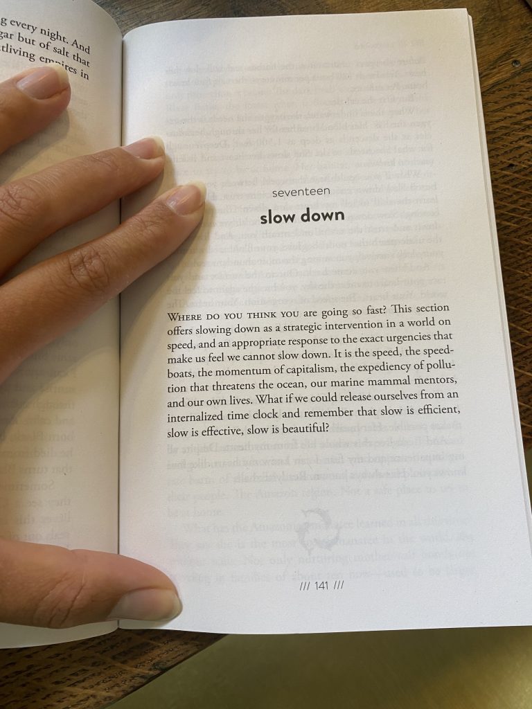 Hand of artist Rebekah Ubuntu holds open Alexis Pauline Gumbs’ book ‘Undrowned: Black Feminist Lessons from Marine Mammals’ on a page titled 'slow down'  