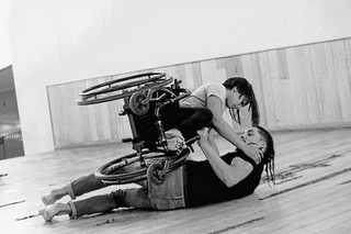 Black and white image of a woman with long dark hair sits horizontally held in her wheelchair in a dance pose. A man lying on the ground holds the woman and her wheelchair above him. 