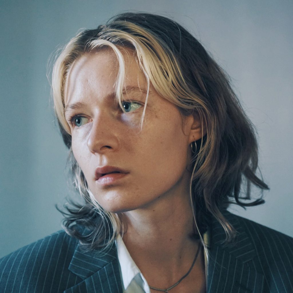 A photographed portrait in cold tones, with white background in the distance. The image is square and shows violet's face and shoulders. It shows a white non-binary person, with shoulder-length hair that is bleached blond at the very front, and the rest of the hair is natural ashy blond. violet has blue eyes and rather small facial features. The expression on their face is neutral, they are looking away from the camera, into the distance. They are wearing an oversized suit jacket that is grey and has thin white stripes, underneath it they are wearing a white shirt opened up, you can see silver jewellery like a small hoop earring and a silver necklace.