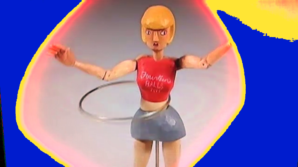 A still from a video. Two superimposed images in bright harsh colours, in the centre of it there is a wooden figurine/doll with a hula hoop on its hips. The figurine has blonde wooden hair in a bob, light skin tone, it is wearing a wooden miniskirt in dusty blue and a red crop top that says BALLS.