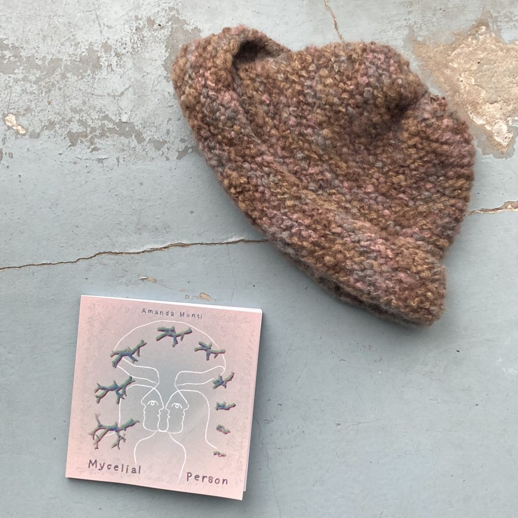 A photograph of two objects lying on a light blue surface: a book of poetry called "Mycelial Person" by Amanda Monti and a knitted winter hat next to it. The pallette of the book cover matches perfectly the colours of the hat. The book cover also has a drawing of a person wearing a mushroom hat, and the winter hat in the picture can be loosely described as mushroom-shaped as well.