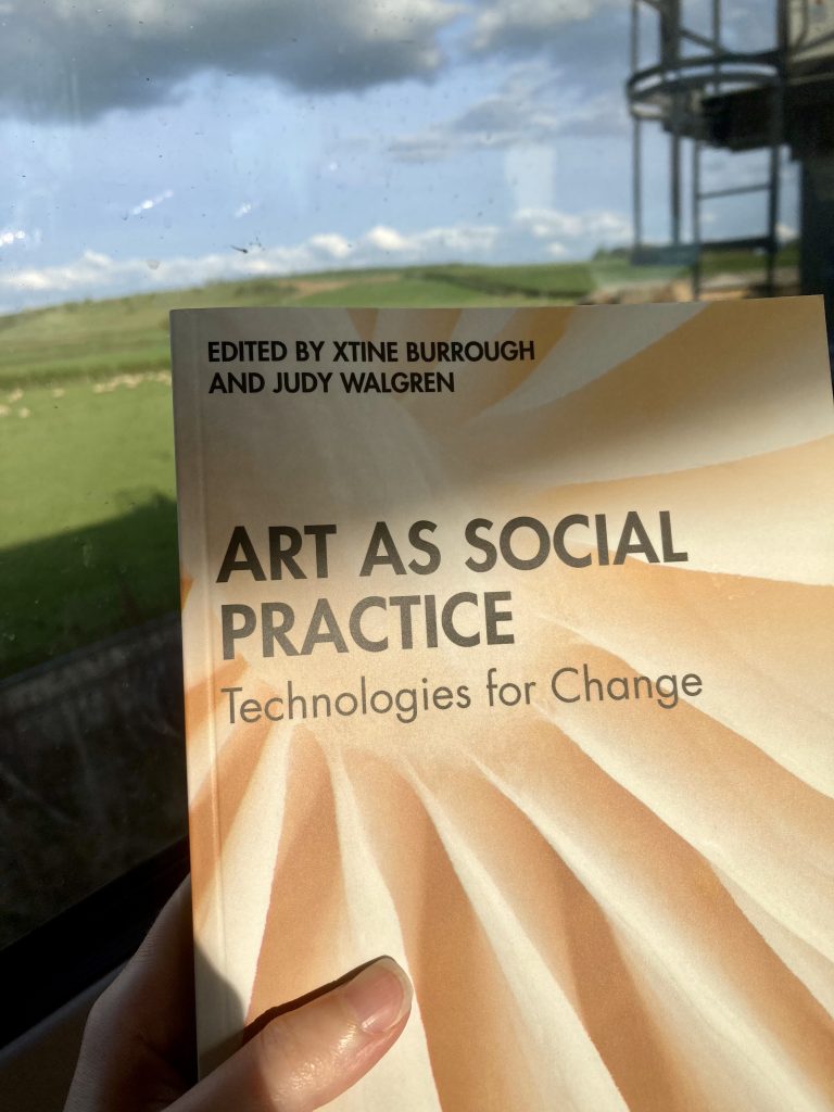 Photograph of the book 'Art as social practice technologies for change' in front of a train window with fields and sheep behind. 